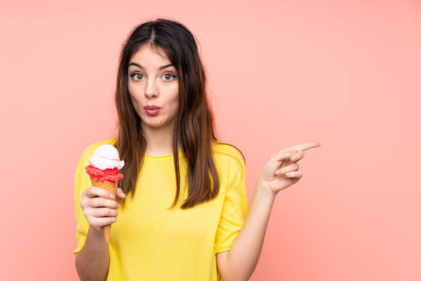 Young brunette woman holding a cornet ice cream over isolated pink background surprised and pointing side - Zdjęcie, obraz