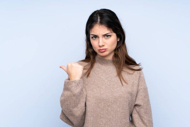 Young brunette woman wearing a sweater over isolated blue background unhappy and pointing to the side - Photo, Image
