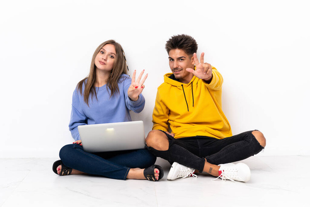Mladý pár s notebookem sedí na podlaze izolované na bílém pozadí šťastný a počítání tři s prsty - Fotografie, Obrázek