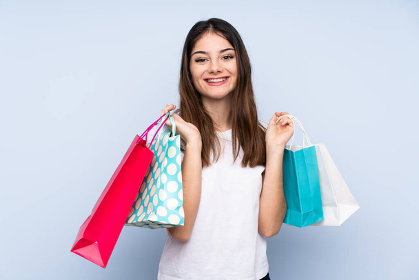 Mladá brunetka žena přes izolované modré pozadí drží nákupní tašky a usmívá - Fotografie, Obrázek