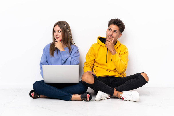 Giovane coppia con un computer portatile seduto sul pavimento isolato su sfondo bianco guardando di lato e sorridente - Foto, immagini