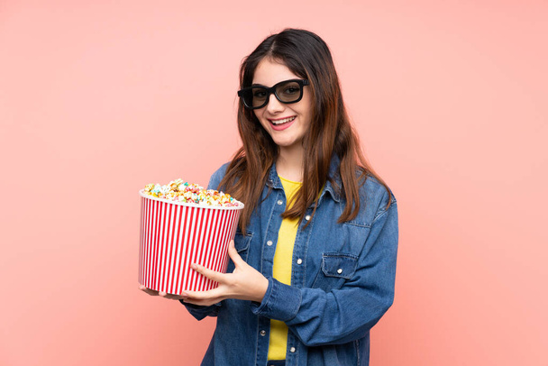Junge brünette Frau auf isoliertem rosa Hintergrund mit 3D-Brille und einem großen Eimer Popcorn - Foto, Bild