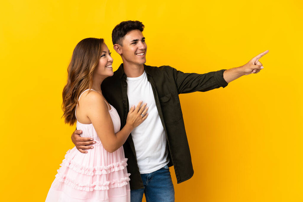 Young caucasian couple isolated on yellow background pointing finger to the side and presenting a product - Фото, изображение