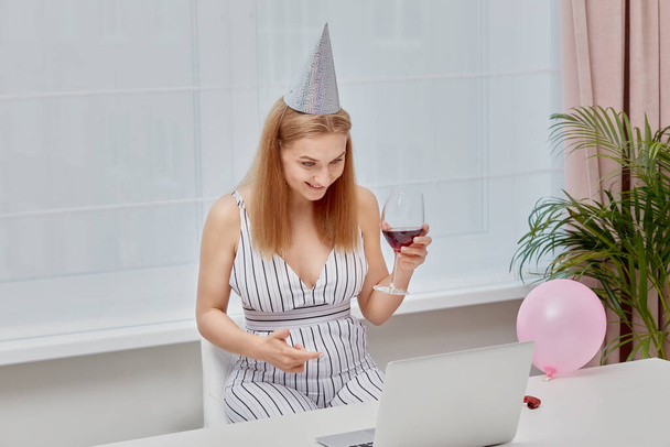Happy blonde girl with glasses of wine celebrates a birthday in front of laptop. - Zdjęcie, obraz