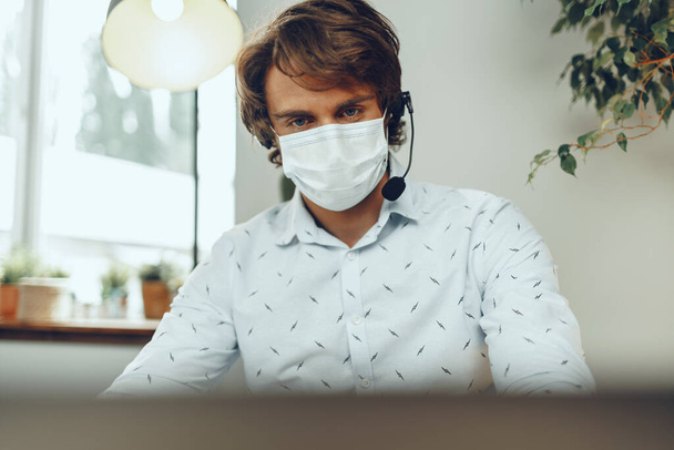 Ο άνθρωπος με την ιατρική μάσκα εργάζεται από το σπίτι ενώ η καραντίνα coronavirus - Φωτογραφία, εικόνα