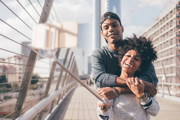Couple noir en plein air prenant selfie à l'aide de bâton selfie - Deiverse homme et femme étreignant en plein air prenant des photos à l'aide d'un smartphone - voyageur, bonheur, concept de convivialité - Photo, image