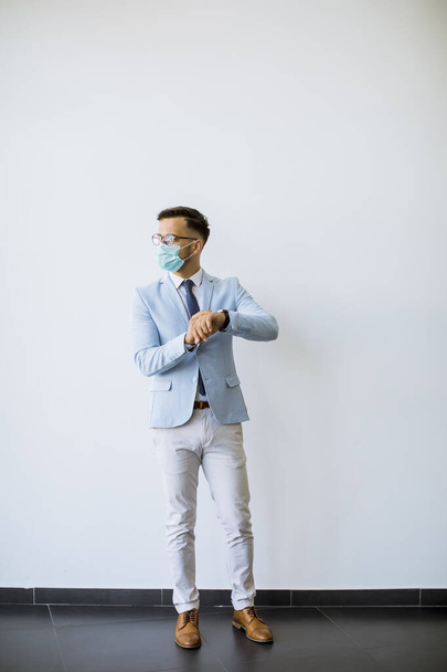 Молодий і стильний бізнесмен з маскою для обличчя стоїть біля стіни в офісі і перевіряє час на годиннику
 - Фото, зображення