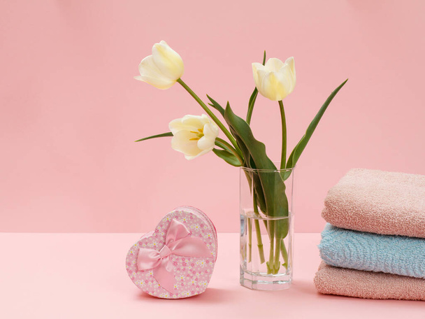 Buquê de tulipas amarelas em um vaso de vidro, uma caixa de presente e toalhas empilhadas em um fundo rosa. - Foto, Imagem