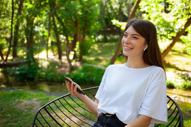 People lifestyle and technology concept. Young funny woman student listen music with wireless headphones, has upbeat mood, enjoys favourite song from playlist, sitting in the park. Empty copy space. - Фото, зображення