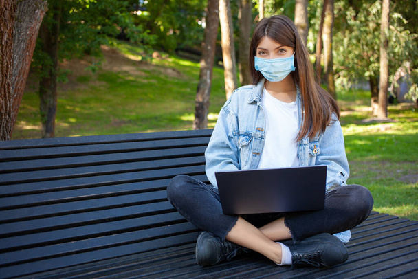 Quarantäne, Fernarbeit und Pandemiekonzept. Eine junge europäische Studentin trägt zum Schutz vor dem Virus eine medizinische Maske, benutzt einen Laptop, sitzt auf der Bank im Park und blickt in die Kamera. Leerer Kopierraum. - Foto, Bild