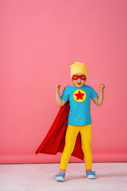 little girl in a superhero costume in a red Cape and hat shows how strong she is. The baby raises her hands in the air, laughs merrily and poses with pleasure in the Studio on a pink background. - Photo, image