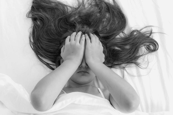 Ein kleines Mädchen liegt im Bett und bedeckt ihr Gesicht mit den Händen. Schwarz-Weiß-Foto. Welttag der Gewaltlosigkeit - Foto, Bild