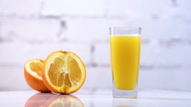 Jugo de naranja recién exprimido en un vaso sobre una mesa con rodajas de naranjas al lado. Refrescante bebida amarilla con frutas sobre fondo blanco - Imágenes, Vídeo