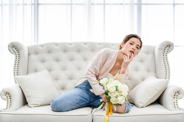 Hermosa mujer asiática encantadora acostada en un sofá blanco y sosteniendo el ramo de rosas blancas sorpresa de su novio en la sala de estar por la mañana. Felicidad relajación estado de ánimo concepto  - Foto, imagen
