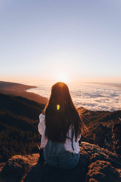 Lang zwart haar meisje kijken naar de zonsondergang van de top van een berg boven een zee van wolken - Foto, afbeelding