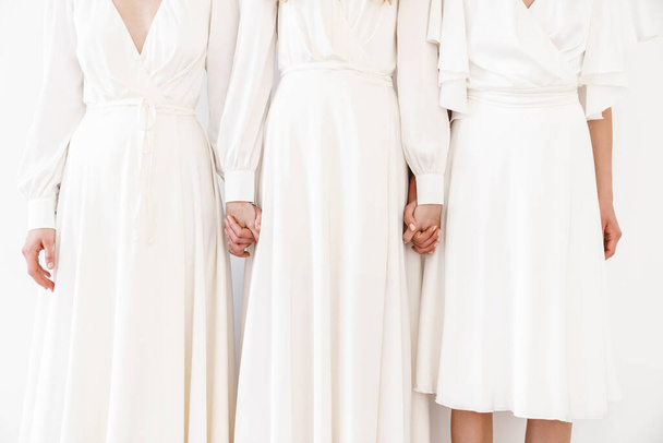 Foto recortada de tres mujeres en vestidos tomados de la mano aislados sobre la pared blanca - Foto, Imagen