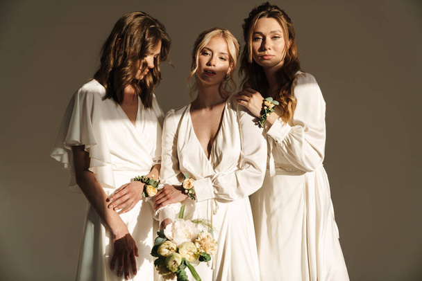 Photo of young beautiful bride with her bridesmaids in white dresses posing with flowers on camera isolated over grey wall - Fotografie, Obrázek