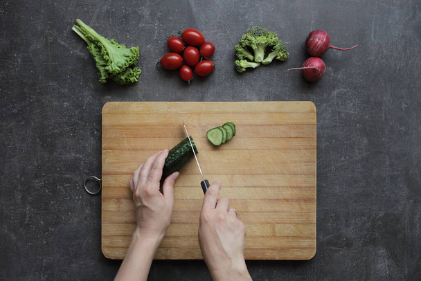 Hands cut cucumber on a cutting board on a gray table. Fresh vegetables, tomatoes, lettuce, broccoli, radishes lie nearby - Photo, Image