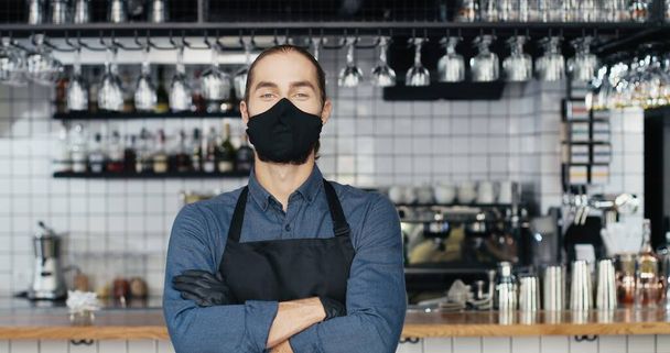 Portrait of Caucasian young handsome man barrista in mask and gloves standing at counter in bar, looking at camera and crossing hands. Waiter posing in cafe with drinks equipment on background. - Photo, Image