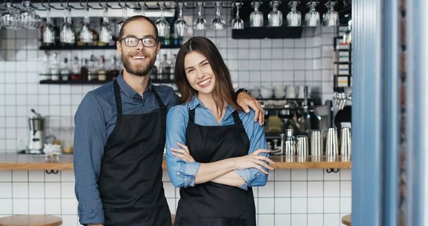 Portrait of Caucasian cheerful couple of bartenders in aprons hugging each other and smiling to camera happily. Happy waiter and waitress in hugs in cafe. Joyful man embracing woman. Barristas. - Zdjęcie, obraz