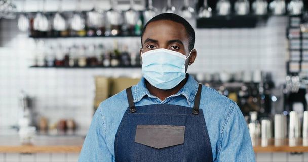 Portrait of African American young handsome man barrista in medical mask standing at counter in bar, looking at camera, crossing hands. Happy waiter smiling in cafe with drinks equipment on background - 写真・画像
