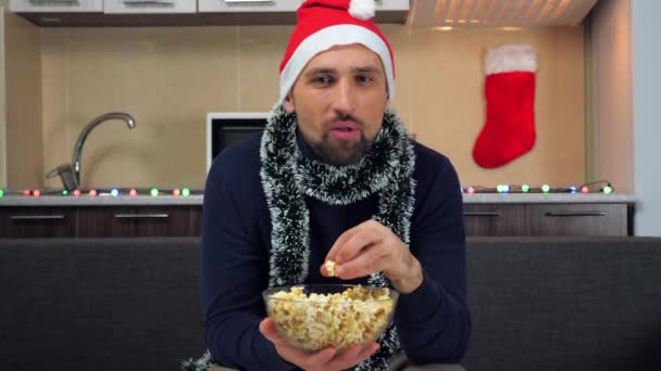 Positieve man Nieuwjaar Kerstman hoed eet popcorn, kijkt tv, lacht, glimlacht - Video