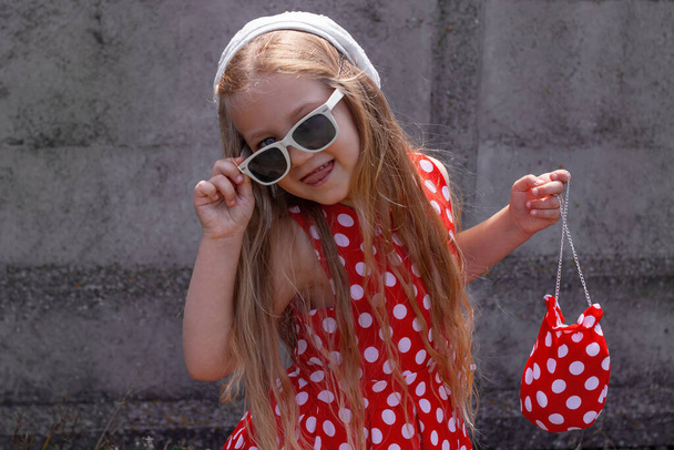 Schöne glückliche Mädchen in einem roten Tupfen-Kleid posiert und lächelt auf Betonmauer Hintergrund. Nettes fröhliches Kind mit langen blonden Haaren, Sonnenbrille und weißem Hut vor grauem Hintergrund draußen. Kleines Modell - Foto, Bild