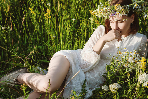 A beautiful woman in a white dress in the boho style with a wreath on her head lies in a field in flowers. The concept of beauty, free life and naturalness - Φωτογραφία, εικόνα