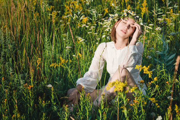 Μια όμορφη γυναίκα με λευκό φόρεμα σε boho στυλ με ένα στεφάνι στο κεφάλι της κάθεται σε ένα χωράφι με λουλούδια. Η έννοια της ομορφιάς, της ελεύθερης ζωής και της φυσικότητας - Φωτογραφία, εικόνα