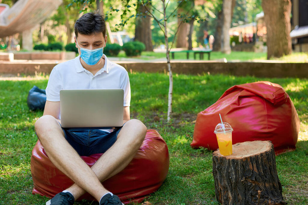 Tıbbi maskeli bir adam parkta bir çanta üzerinde oturuyor ve bir dizüstü bilgisayar üzerinde çalışıyor, Moldova Chisinau yakınlarındaki bir kütükte limonata içiyor. - Fotoğraf, Görsel