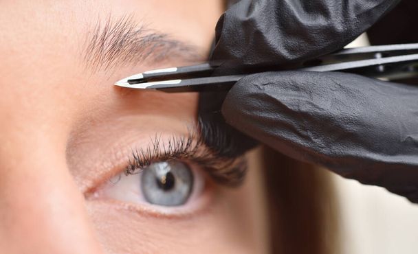 Eye and eyebrow of a young woman and tweezers, eyebrow correction procedure - Foto, afbeelding