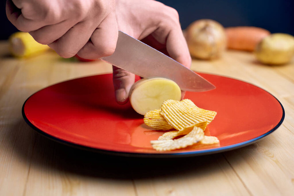 Ένας άντρας να κόβει πατάτες σε πατατάκια σε κόκκινο πιάτο και ξύλινο τραπέζι, λαχανικά τριγύρω, ιδέες για πρόχειρο φαγητό. - Φωτογραφία, εικόνα