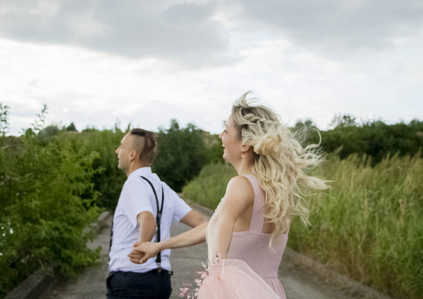 Casamento criativo hipster à beira-mar. A noiva está em um vestido rosa e véu, o noivo está em belas calças com suspensórios e uma camisa de juventude. Loucura, diversão e uma abordagem incomum ao casamento
 - Foto, Imagem