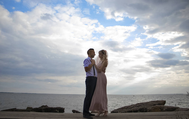 Kreative Hipster-Hochzeit am Meer. Die Braut trägt ein rosafarbenes Kleid und einen Schleier, der Bräutigam eine schöne Hose mit Hosenträgern und ein Jugendhemd. Wahnsinn, Spaß und ein ungewöhnlicher Umgang mit der Ehe - Foto, Bild