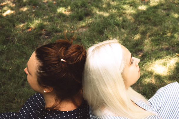 Δύο νεαρές γυναίκες κάθονται πλάτη με πλάτη στο γρασίδι. Κλείσε. Πλευρική άποψη - Φωτογραφία, εικόνα