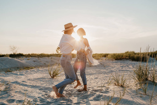 sunny two young women having fun on the sunset beach, queer non-binary gender identity, gay lesbian love romance, boho summer vacation style wearing jeans - 写真・画像