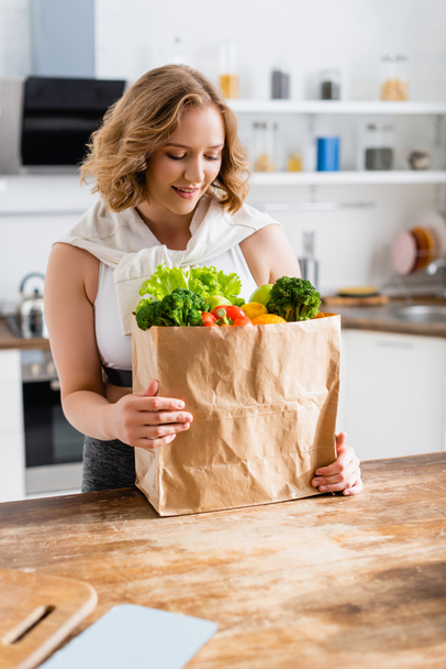 young woman looking at paper bag with groceries in kitchen  - 写真・画像