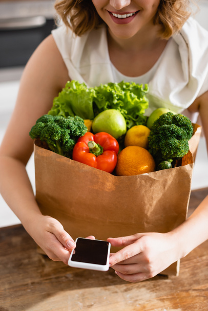cropped view of woman holding smartphone with blank screen near groceries  - Foto, afbeelding