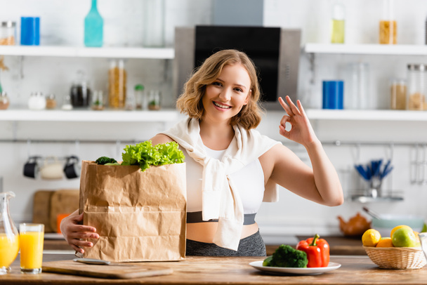 young woman showing ok sign near paper bag with lettuce and bowl with fruits - Foto, afbeelding