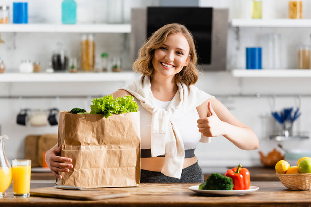 young woman showing thumb up near paper bag with lettuce and bowl with fruits - Photo, Image