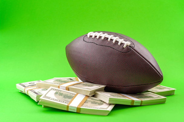 Winning bet on sporting event, money in sport and sports betting conceptual idea with american football ball and wads of cash isolated on green background - Photo, Image