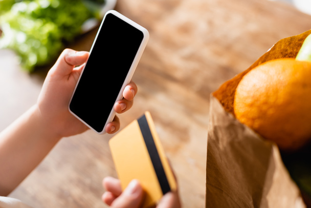 selective focus of woman holding smartphone with blank screen and credit card near fruits in paper bag  - Foto, afbeelding