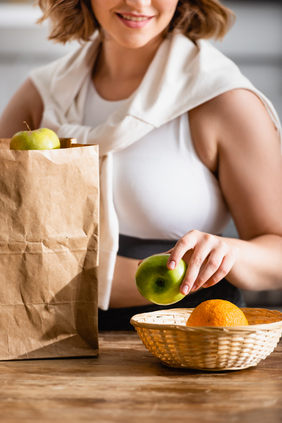 partial view of woman holding apple near paper bag and fruits  - Фото, изображение