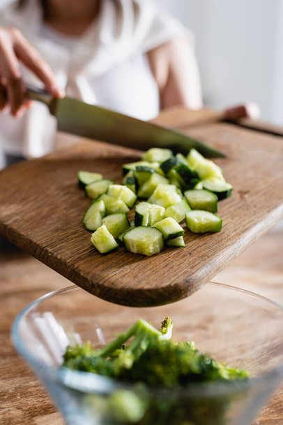 cropped view of woman holding cutting board with sliced cucumber near bowl with fresh broccoli  - Fotografie, Obrázek