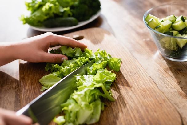 selective focus of woman cutting green lettuce on chopping board near sliced cucumber in bowl   - Fotografie, Obrázek