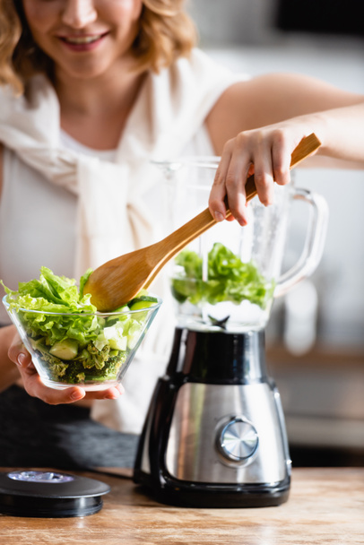 cropped view of woman holding bowl with fresh lettuce and wooden spoon near blender  - Fotografie, Obrázek