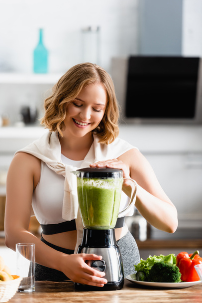 young woman mixing green smoothie in blender near vegetables  - Fotografie, Obrázek