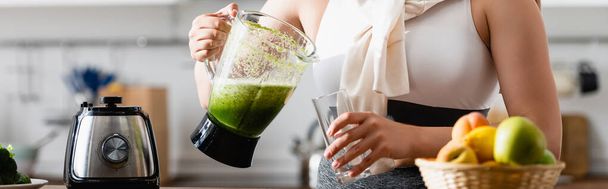 panoramic crop of young woman holding blender with mixed smoothie near glass  - Photo, Image