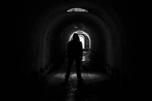 Kapüşonlu yalnız bir adam, Vladivostok, Fort Pospelova 'daki karanlık ve terk edilmiş bir yeraltı koridorundan aydınlığa doğru yürür.. - Fotoğraf, Görsel