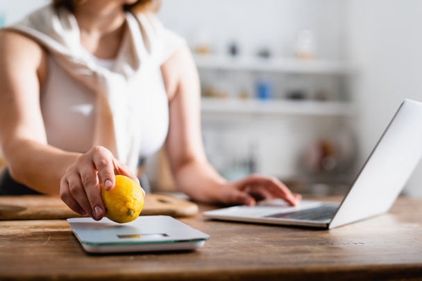 bijgesneden weergave van vrouw zetten citroen op keukenweegschalen en met behulp van laptop  - Foto, afbeelding